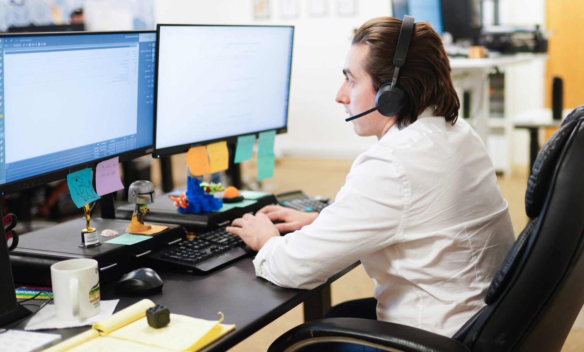 A young man typing on a computer at his desk. There are various sticky notes on his monitor and he is wearing a headset.